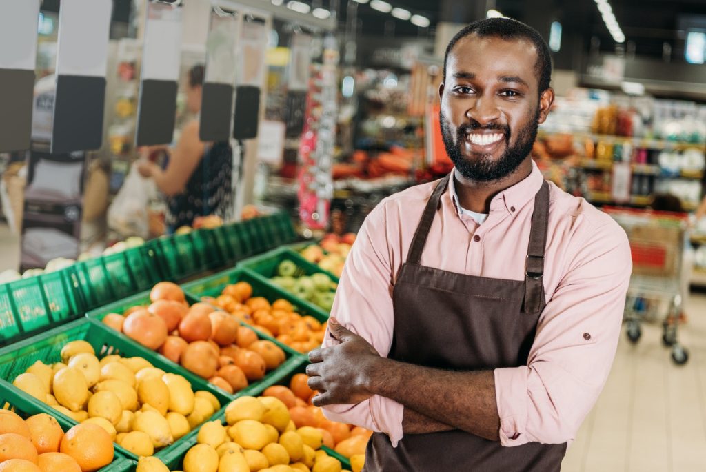 african american male shop assistant in apron standing with crossed arms near fruit department in